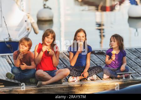 Eine Gruppe von vier Kindern, 3 Mädchen und ein Junge, die im Freien Eiscreme essen und sich auf einem Pier am See ausruhen Stockfoto