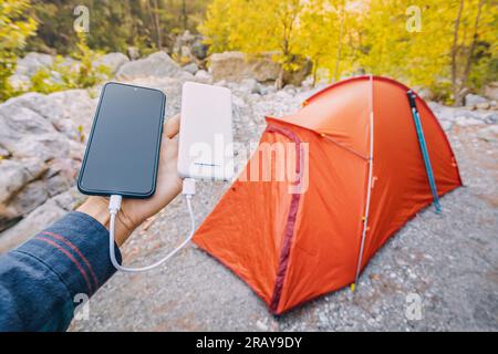 Powerbank zum Aufladen von Smartphones und anderen elektrischen Geräten am Wochenende bei Wanderungen. Orangefarbenes Campingzelt im Hintergrund Stockfoto