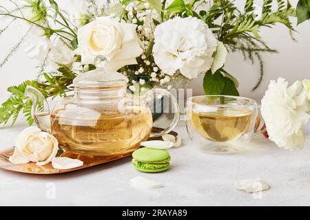 Kräuterteekanne, Tasse, Makronen-Dessert, Lebensstil mit weißen Blumen. Ästhetisch-feminin, meditative Zeit für sich selbst, gemütliches Zuhause Stockfoto