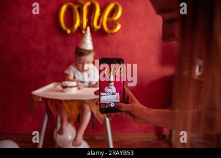 Die Hand der Mutter macht Fotos von ihrem Sohn auf einem Smartphone. Ein einjähriger Junge mit festlichem Hut, der Geburtstagskuchen mit einer Kerze in den Händen hält. Auf der roten Ebene Stockfoto