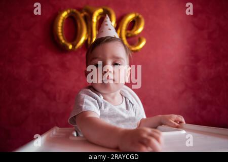 Portrait eines einjährigen Jungen mit festlichem Hut sitzt auf einem Kinderstuhl mit Stofffahnen. Kind auf isoliertem roten Hintergrund mit goldenen Luftballons aus Folie Stockfoto