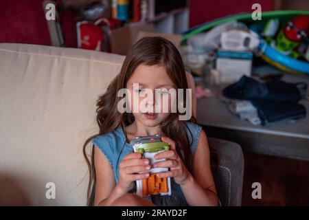 Das Porträt eines kleinen Mädchens mit langen Haaren sitzt zu Hause auf dem Sofa. Das Mädchen hält Spielzeug in den Händen und erzählt etwas. Innenraum mit Chaos im C Stockfoto