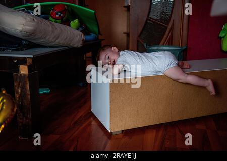 Porträt eines Kleinkindes im grauen Bodysuit, das auf einer weißen Kommode in einem Spielzeugchaos im Kinderzimmer liegt. Erziehung in Kreativität, Handlungsfreiheit. Ba Stockfoto