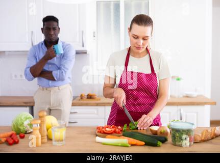 Frau bereitet Gemüsesalat in der Küche zu, während ihr Mann aus einem Kaffeebecher trinkt Stockfoto