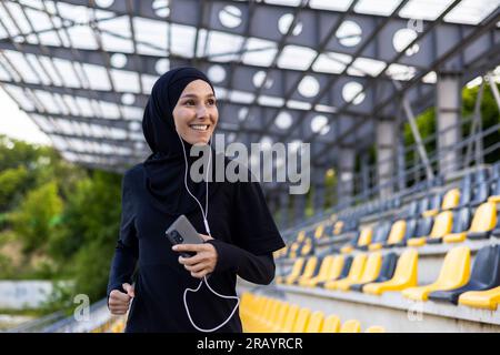 Junge, wunderschöne muslimische Frau, die im Stadion im Hijab läuft, eine Sportlerin, die in Kopfhörern lächelt, ein Telefon hält und Hörbuch hört. Stockfoto