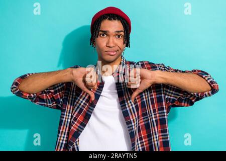 Foto eines unzufriedenen, enttäuschten Mannes mit Dreadlocks, das ein kariertes Hemd trägt, das die Daumen nach unten zeigt, isoliert auf türkisfarbenem Hintergrund Stockfoto