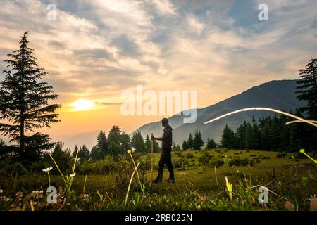 28. Juni 2023, Nagthat, Uttarakhand, Indien: Trekker mit Stock in Wiese bei Sonnenuntergang, umgeben von Bergkämmen und Kiefern. Himalaya, Uttarakhan Stockfoto