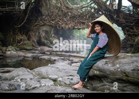 Ein Mädchen aus dem khasi-Stamm in traditioneller Kleidung mit traditionellem Regenschutz aus Bambus, sitzt vor der lebenden Wurzelbrücke, Meghalaya, Indien Stockfoto