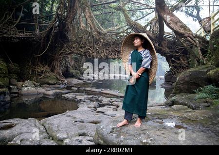 Junge Frau mit traditioneller Kopfregenabdeckung, die am Fluss vor einer lebenden Wurzelbrücke steht, Meghalaya, Indien Stockfoto