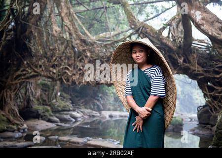 Junge Frau mit traditioneller Kopfregenabdeckung, die am Fluss vor einer lebenden Wurzelbrücke steht, Meghalaya, Indien Stockfoto