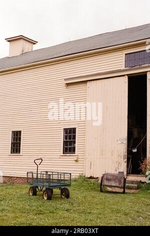 Newbury, Massachusetts: Im historischen Spencer Pierce Littl befindet Sich Ein grüner Gartenwagen aus Metall vor einer großen beigefarbenen Milchscheune aus Holz und grünem Gras Stockfoto