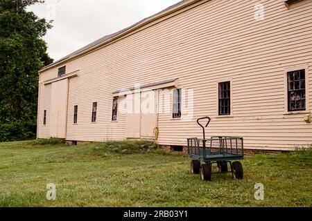 Newbury, Massachusetts: Im historischen Spencer Pierce Littl befindet Sich Ein grüner Gartenwagen aus Metall vor einer großen beigefarbenen Milchscheune aus Holz und grünem Gras Stockfoto