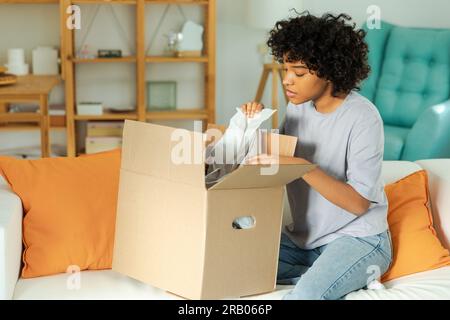 Afrikanisches Mädchen, das die Lieferung auspackt und in die Schachtel schaut. Glückliche Frau, die eine Schachtel öffnet. Eine Frau bekommt ein Paket, das sich die gelieferten Waren ansieht. Zufriedene Kunden positives Feedback, schnelle Expresslieferung Stockfoto