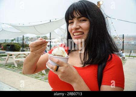Die junge venezolanische Latina-Frau lächelt fröhlich in die Kamera, während sie eine kleine Tasse Vanille und Schokoladeneis mit einem Plastikspoo isst Stockfoto