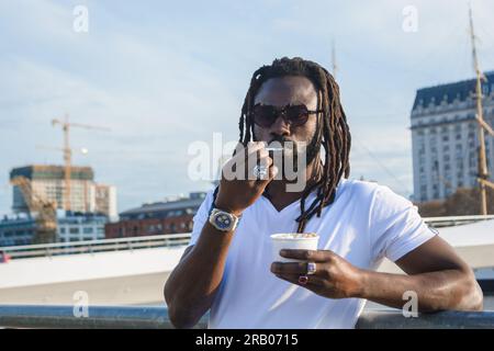 Porträt eines jungen schwarzen ethnischen Mannes mit Dreadlocks, Bart und Sonnenbrille, er ruht sich aus und isst ein Eis und genießt das Wochenende in Buenos Aires Stockfoto