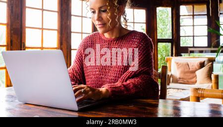 Eine Frau mittleren Alters, die zu Hause einen Computer benutzt, mit heiterem Gesichtsausdruck und rotem Pullover. Holzhüttenhaus. Alternative Bürojobs für den Tischarbeitsplatz. Leute, die allein im Netz surfen. Reisen Stockfoto