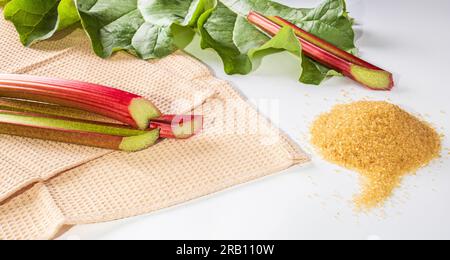 Gesundes Essen. Rhabarberstiele auf Waffeltuchhintergrund, Rhabarberblätter und ein Haufen brauner Zucker auf weißem Hintergrund Stockfoto
