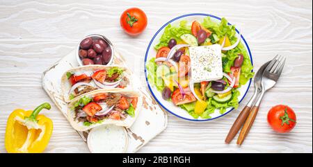 Traditionelle griechische Speisen: Griechischer Salat, Gyros mit Fleisch und Gemüse, Tzatziki-Sauce, Oliven auf weißem rustikalem Holztisch von oben Stockfoto