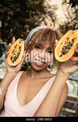 Fröhliche junge afroamerikanische Frau mit Zahnspangen, Sommerkleid und Kopftuch, während sie geschnittene Papaya in der Hand hält und in Orangerie, fashi, in die Kamera schaut Stockfoto