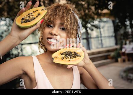 Porträt einer positiven jungen afroamerikanischen Frau mit einer Zahnspange, die Sommerkleidung trägt und frische Papaya hält, während sie in einer orangery, trendigen Frau steht Stockfoto