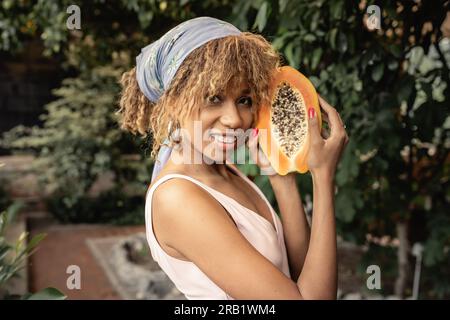 Eine junge afroamerikanische Frau mit einer Zahnspange, einem Sommerkleid und einem Kopftuch mit frischer Papaya und einem Blick auf die Kamera in Orangerie, stilvoller La Stockfoto