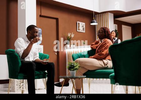 Hotelgäste sitzen im Loungebereich des modernen Resorts und warten auf den Check-in an der Rezeption. Ein afroamerikanisches Paar, das in der Hotellobby unterkommt, und Mitarbeiter, die exzellenten Service bieten. Stockfoto