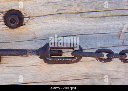 Kettenglieder sind mit einem großen rostigen Bolzen miteinander verbunden, wodurch eine feste Verbindung entsteht. Die Kette dehnt sich über verwittertes Holz. Stockfoto