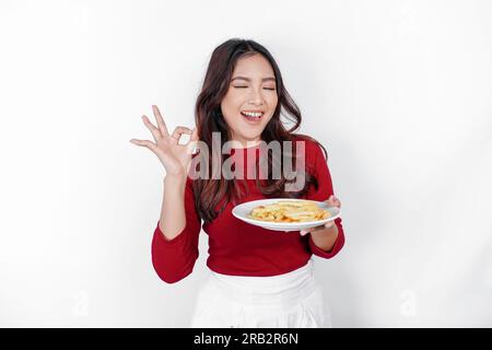 Bild eines lächelnden jungen asiatischen Mädchens, das einen Teller pommes frites in der Hand hält und das OK-Schild zeigt, isoliert auf weißem Hintergrund Stockfoto