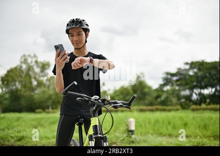 Ein attraktiver und fitter junger asiatischer Mann in Sportbekleidung und ein Fahrradhelm sitzt auf seinem Fahrrad, benutzt sein Smartphone und schaut auf seine Smartwatch. Stockfoto