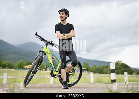 Ein Porträt eines gesunden, fitten jungen Asiaten in Sportbekleidung und ein Fahrradhelm stehen mit den Armen auf der Straße mit seinem Fahrrad. Stockfoto