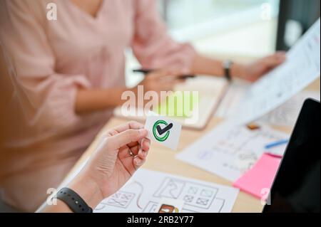 Nahaufnahme einer weiblichen UI UX-Entwicklerin, die während des Meetings mit ihrem Team ein Papier mit einem korrekten Schild in der Hand hielt. Richtig, richtig, ja, genehmigen, zustimmen Stockfoto