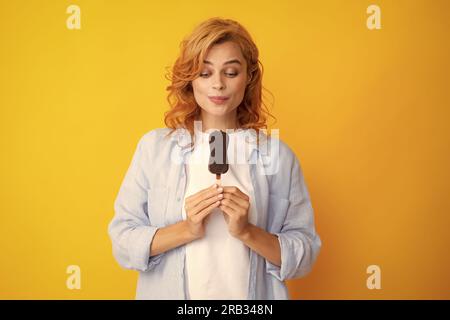 Junge Frau isst leckeres Eis, genießt gefrorenes Dessert, in legerer Kleidung gekleidet, isoliert auf gelbem Hintergrund. Stockfoto