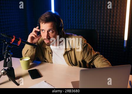 Mann in Senfhemd mit Kopfhörern, der am Mikrofon Gesten trägt und seine Geschichte mit dem Publikum teilt, während er im Studio mit Neonbeleuchtung am Schreibtisch sitzt Stockfoto