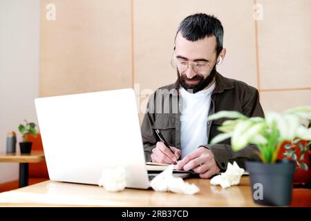 Konzentrierter Mann mit Brille, der sich Notizen macht, Informationen aus einem Laptop in einem Café schreibt, ernster bärtiger Mann, der im Café lernt Stockfoto