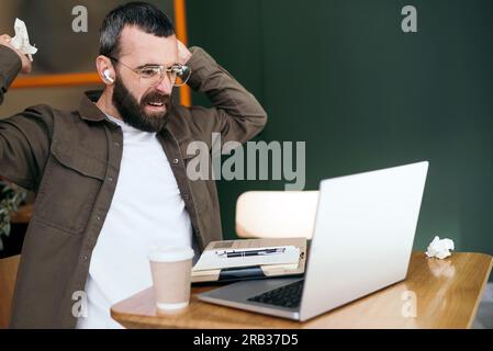 Wütender, bärtiger Freiberufler im Café, der sich streitet, über Webcam spricht, Probleme mit Online-Geschäftsprojekten diskutiert, Notizbuchseiten reibt Stockfoto