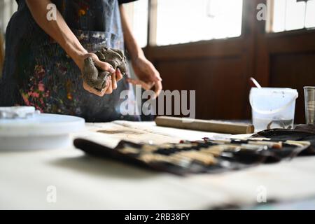 Töpferkünstler in Schürze kneten ein Stück rohen Ton mit den Händen auf einem Holztisch. Handwerk, Kreativität, Hobby und Aktivitätskonzept Stockfoto