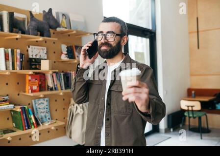 Ein stylischer Mann mit Bart und Brille steht auf einem Buchhintergrund, hält eine Tasse Kaffee in einem Café und lächelt in die Kamera. Stockfoto