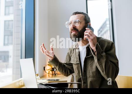 Wütender weißer Freiberufler im Café, der Streit während eines Telefongesprächs hat und Probleme mit einem Online-Geschäftsprojekt diskutiert Stockfoto