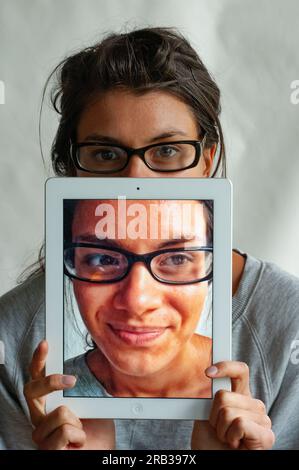 Eine junge Frau, die ihren iPadto benutzt, versteckt sich dahinter, auf dem ein Foto von ihrem Gesicht ist Stockfoto