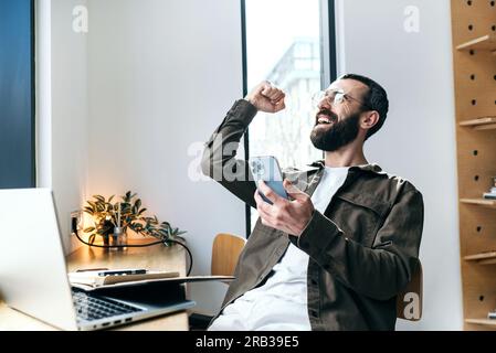 Ein fröhlicher bärtiger Mann mit Brille jubelt in einem Café mit einem Handy und macht eine Siegergeste. Ferne Arbeit. Café-Bar, Fernarbeiten, Freiberufler Stockfoto