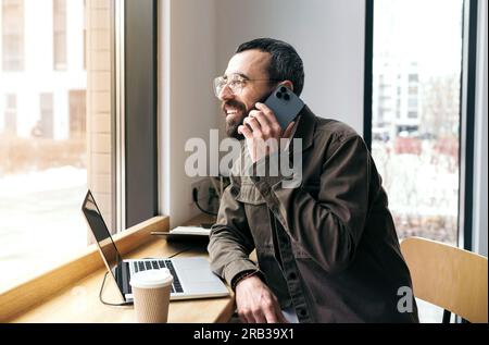 Ein Weißer spricht in einem Café mit einem Handy. Arbeitsprozess Ein Mann mit Bart im Hemd sitzt in einem Café mit einem Laptop und einem Telefon. Stockfoto