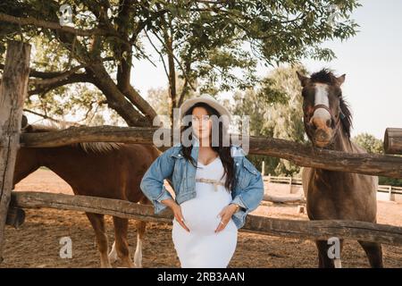 Stilvolle schwangere Frau mit weißem Hut auf dem Land in der Nähe eines Pferdes. Stockfoto