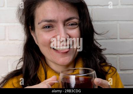 Eine hübsche weiße Frau mittleren Alters trinkt Tee und lacht verspielt, indem sie ihre Zunge zeigt, während sie Zeit mit Freunden verbringt. Transparenter Becher mit Feinkost Stockfoto