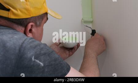 Rückansicht eines Mannes in einem grauen T-Shirt und einer grauen Kappe, der die Wand mit einer grünen Malerwalze mit grüner Farbe lackiert. Ein Mann malt mit einem Malerwalzen eine Wand. Tun Stockfoto