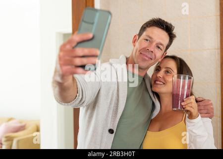 Glückliches weißliches Paar, das einen Fruchtcocktail trinkt und Selfie mit dem Smartphone in der Küche macht. Gesunder Lebensstil, Essen und häusliches Leben, unverändert. Stockfoto