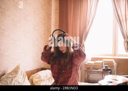 Junge Frau, die zu Hause auf dem Bett sitzt und Kopfhörer aufsetzt Stockfoto