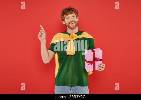 Feier, fröhlicher rothaariger Mann mit Bart in lässiger Kleidung auf Korallenhintergrund, mit Geschenkbox, festlichen Anlässen, Geschenk, Mode und Trend Stockfoto