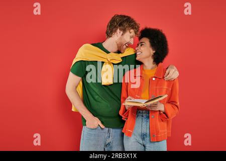 Interrassisches Paar, das sich umarmt und ein Buch über Korallenhintergrund, kulturelle Vielfalt, lebendige Farben, stilvolles Outfit, Jugend und Intelligenz liest, Multi-Parameter Stockfoto