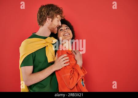 Interrassisches Paar, das sich mit Korallen umarmt und lächelt, kulturelle Vielfalt, lebendige Farben, stilvolle Outfits, Jugend und Mode, Gemischtrassige Menschen Stockfoto