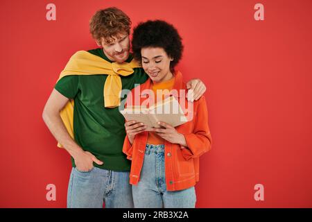 Interrassisches Paar, das sich umarmt und ein Buch über Korallen, kulturelle Vielfalt, lebendige Farben, stilvolle Outfits, Jugend und Intelligenz liest, Multikulär Stockfoto